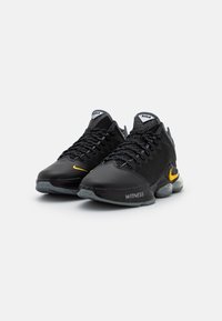 Le scarpe sportive nere presentano una parte superiore testurizzata, dettagli in rete e un logo giallo ben visibile sul lato. Suola in gomma trasparente.
