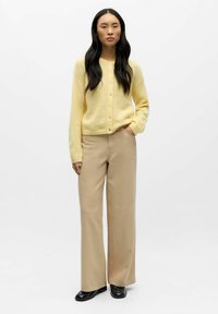 Dzeltena adīta kardigans ar pogu aizdari, kopā ar plašām bēšām biksēm un melniem apaviem. Vienkāršs dizains, mīksta tekstūra un neitrālas krāsas.