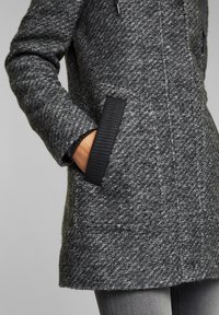 Main dans la poche d’un manteau gris foncé texturé avec un bord côtelé noir sur la poche et un pantalon gris visible sous le manteau.