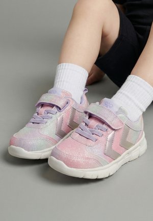 Dziecko noszące błyszczące różowo-fioletowe trampki na rzepy i białe skarpetki, siedzące na szarej podłodze.