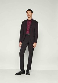 Jovem em pé, vestindo um fato preto, uma camisa bordô abotoada, sapatos de vestir pretos, contra um fundo cinza claro liso.