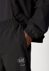 Vêtements de sport noirs avec une poche zippée et des poignets élastiques. Comprend un détail de logo blanc avec "EA7" et "VENTUS7" sur le pantalon.