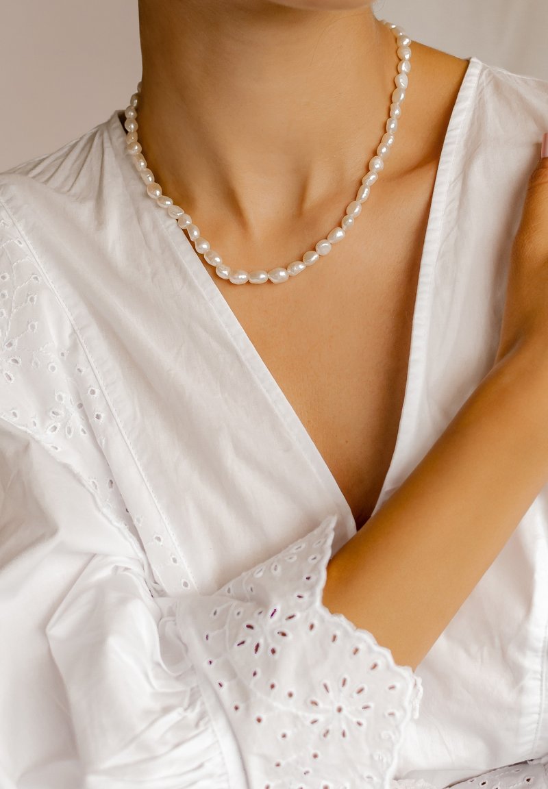 Collar de perlas blancas con cuentas ovaladas de diferentes tamaños, llevado sobre un cuello desnudo, con una blusa blanca texturizada que presenta detalles de ojal.
