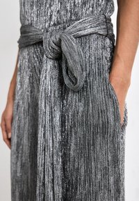 Sølvmetallic jumpsuit med bælte i taljen. Har vertikale plisseringer, en glat tekstur og sidelommer.