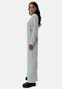 Abito maglione lungo a coste di colore grigio chiaro, realizzato in un morbido tessuto a maglia. Presenta maniche ampie e una silhouette dritta. Scarpe con plateau nere.