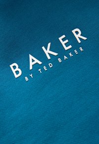 Tegen een teal-kleurige stof met een gestructureerd oppervlak staat het woord "BAKER" in witte, vetgedrukte letters boven "by TED BAKER," duidelijk leesbaar.