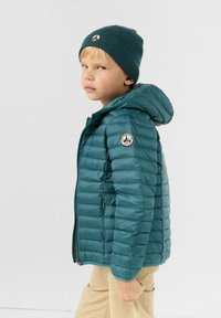 Groenejas met capuchon, voorzien van horizontale doorgestikte lijnen en een logo op de mouw. Gecombineerd met een bijpassende geribbelde muts.