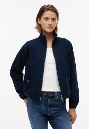 Rövid barna hajú nő, aki sötétkék cipzáras dzsekit visel fehér felső és kék farmernadrág felett, egyik kezét a zsebében tartva áll.