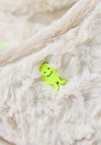 Una tela beige difusa con una textura suave presenta una pequeña figura sonriente de plástico verde incrustada en ella.