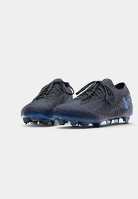Scarpe da calcio nere e blu con tomaia texturizzata, lacci piatti e tacchetti conici per la presa. Presentano un logo Under Armour angolare.