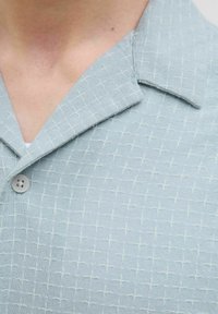 Camisa de manga corta azul claro con una tela texturizada, que presenta un patrón de cruces y un cuello abotonado.