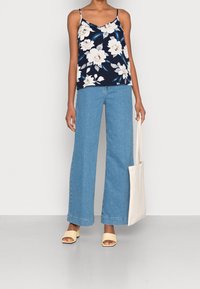 Camisole floreale navy con spalline sottili, abbinato a jeans a vita alta di colore blu chiaro e a gamba larga. Il modello tiene una tote beige e indossa sandali beige.