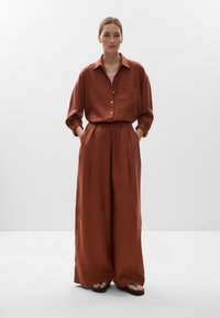 Camicetta di lino color ruggine con chiusura a bottoni e taschino sul petto, abbinata a pantaloni larghi coordinati. Il modello indossa sandali piatti.