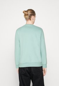 Person med blont hår i en bun, iklädd en ljusgrön långärmad sweatshirt och svarta kordjaktsbyxor, vänd bort från kameran mot en vit bakgrund.