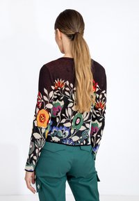Pull noir avec une broderie florale colorée en rouge, jaune, bleu et vert. Porté sur un pantalon bleu-vert, mettant en valeur une coupe ajustée.