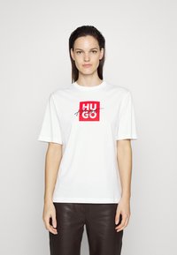 Biely bavlnený tričko so stredovou červenou štvrcovou grafikou a nápisom "HUGO" v čiernej kurzívou. Krátke rukávy, okrúhly výstrih, voľný strih.
