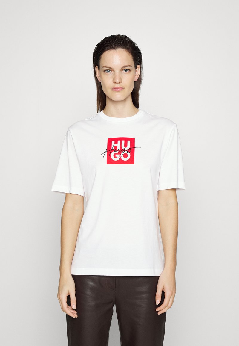 Biely bavlnený tričko so stredovou červenou štvrcovou grafikou a nápisom "HUGO" v čiernej kurzívou. Krátke rukávy, okrúhly výstrih, voľný strih.
