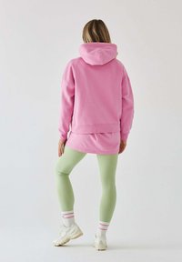 Różowa bluza z kapturem o luźnym kroju, nałożona na dłuższą koszulę, zestawiona z pastelowymi zielonymi legginsami i białymi sneakersami z różowymi akcentami.