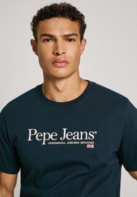 Tamsiai mėlynos spalvos medvilninė marškinėliai, kuriuose matosi ryškus baltas logotipas "Pepe Jeans" ir tekstas "ORIGINAL DENIM BRAND" apačioje, su mažyte Jungtinės Karalystės vėliavos detalė.