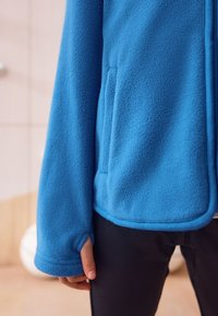 Jachetă albastră din fleece cu textură moale, prevăzută cu buzunare laterale și croială lejeră. Mânșetele sunt ușor prelungite.