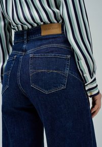 Jean en denim bleu foncé avec une étiquette en cuir "Salsa Jeans" sur la ceinture, présentant deux poches arrière avec des détails de couture courbés.
