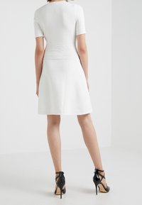 Robe blanche à mi-mollets avec manches courtes, en tissu texturé, et corsage ajusté. Portée avec des chaussures à talons hauts noires avec des brides à la cheville.