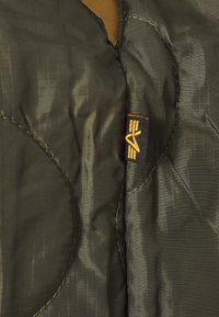 Tissu matelassé vert olive avec une texture lisse, présentant une étiquette noire avec un logo jaune et des détails de couture orange.