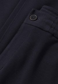 Navy stof met een gladde textuur, met een voorzak met een zwarte knop. Eenvoudig ontwerp met stiksels op de stof.