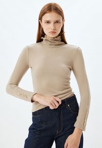 Maglia beige a maniche lunghe con colletto alto arricciato e bottoni decorativi sulle maniche. Tessuto liscio, design fitted, abbinata a jeans scuri.