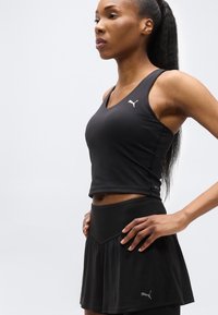 Γυναίκα που φοράει μαύρο αθλητικό τοπ και φούστα Puma, με τα χέρια στη μέση, κοιτάζοντας πλαγίως προς ένα απλό ανοιχτόχρωμο φόντο.