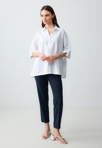 Camicia bianca a maniche corte, morbida e con bottoni, abbinata a pantaloni blu scuro su misura e sandali bianchi con tacco quadrato e fasce.
