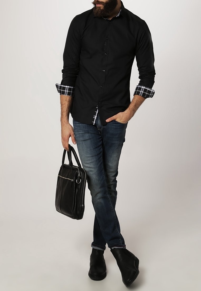 Man with beard wearing black shirt with rolled plaid cuffs, blue jeans, black shoes, holding black leather briefcase, standing with crossed legs.