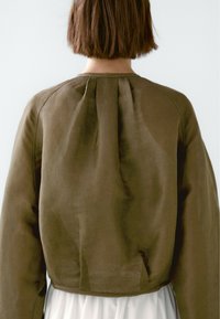Giacca cropped color khaki realizzata in un tessuto leggero e strutturato. Collo rotondo, pieghe sottili sul retro e maniche di lunghezza polso.