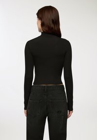 Kobieta z długimi brązowymi włosami, ubrana w czarny, prążkowany top z długimi rękawami oraz wysokim stanem, w jasnych czarnych dżinsach, zwrócona plecami na białym tle.