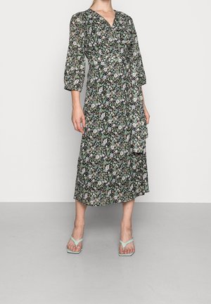 Femme portant une robe portefeuille midi noire à motifs floraux avec manches trois-quarts et sandales à talons ouvertes vert clair.