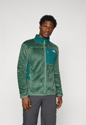 Veste en polaire verte avec une surface texturée, col montant, poche zippée à l'avant et détails de fermeture éclair et accents bleus contrastants.