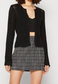 Czarny dzianinowy kardigan z otwartym przodem, misternym wzorem, długimi rękawami i krótkim krojem, zestawiony z wysokim stanem czarno-białą spódnicą w kratę.