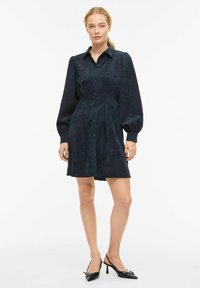 Haljina košulja od tkanine kord s dubokom mornarsko plavom bojom, s dugim rukavima na naduvavanje, prednjim gumbima i strukom prilagođenim tijelu, s naboranim suknjom i glatkom teksturom.