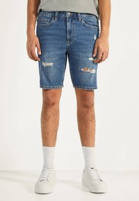 Man som bär blå trasiga denimshorts, vita strumpor och vita sneakers, stående på en vit yta mot en neutral bakgrund.