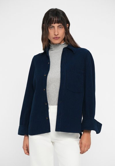 Modra kordna srajca z gumbi, žepom na prsih in dolgimi rokavi. Nosi se preko svetle sive turtlenecka. Bele hlače dopolnjujejo obleko.