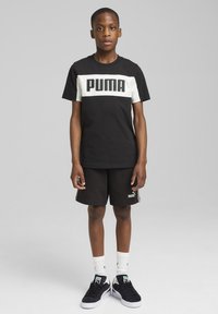 Czarna bawełniana koszulka z białym paskiem i logo "PUMA", zestawiona z czarnymi szortami. Model nosi białe skarpetki i czarne trampki.