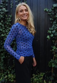 Kék, átlátszó hatású hosszú ujjú felső fekete szívmintákkal. Fekete nadrággal párosítva, sötét fa háttér előtt.