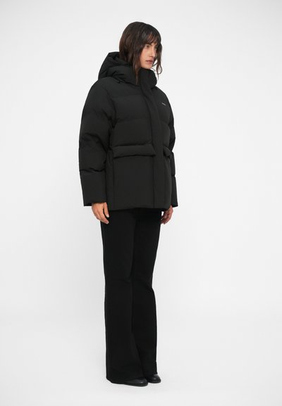 Veste doudoune noire avec capuche, au design ample, texture matelassée et deux poches à l'avant. Porté avec un pantalon noir évasé.