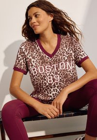 Leopardmönstrad t-shirt med vinröd V-ringning, kantade ärmar och "BOSTON 89"-grafik. Modellen bär matchande vinröda leggings och sitter ned.