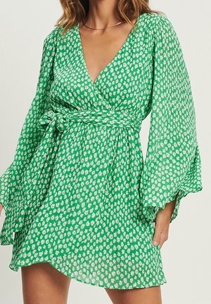 Mujer que lleva un vestido cruzado verde de manga larga con un estampado floral blanco y un cinturón anudado en la cintura.