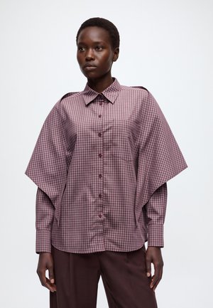 Mannequin portant une chemise mauve à carreaux boutonnée avec des manches cape dramatiques et un pantalon foncé, debout devant un fond clair uni.