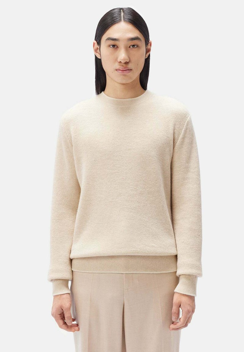 Beige texturerad tröja med rund halsringning och ribbade manschetter. Mjuk tyg, avslappnad passform, bärs över ljusbeiga byxor. Enkel, minimalistisk design.