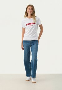Witte katoenen T-shirt met rode tekst "UNWIND again", gecombineerd met blauwe spijkerbroek met flared pijpen en witte sneakers met beige zolen.