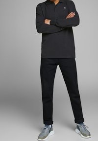 Svart långärmad pikétröja med krage och logodetalj, kombinerad med mörkblå jeans och grå sneakers med blå detaljer.