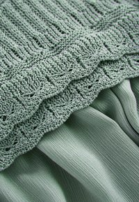 Gros plan sur un tissu vert clair à motif tricoté lâche superposé à un tissu lisse, mettant en valeur les détails textiles et les plis.